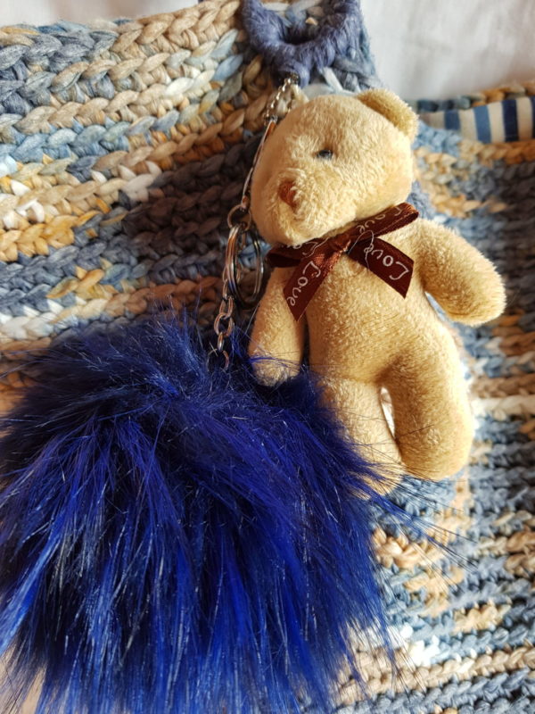 Detalle del bolso hecho a mano con hilo de algodón de mezcla de colores azules y tostados, forrado con tela de loneta rallada y complemento decorativo de oso con pompón azul, asas de piel