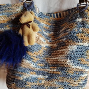 Bolso hecho a mano con hilo de algodón de mezcla de colores azules y tostados, forrado con tela de loneta rallada y complemento decorativo de oso con pompón azul, asas de piel